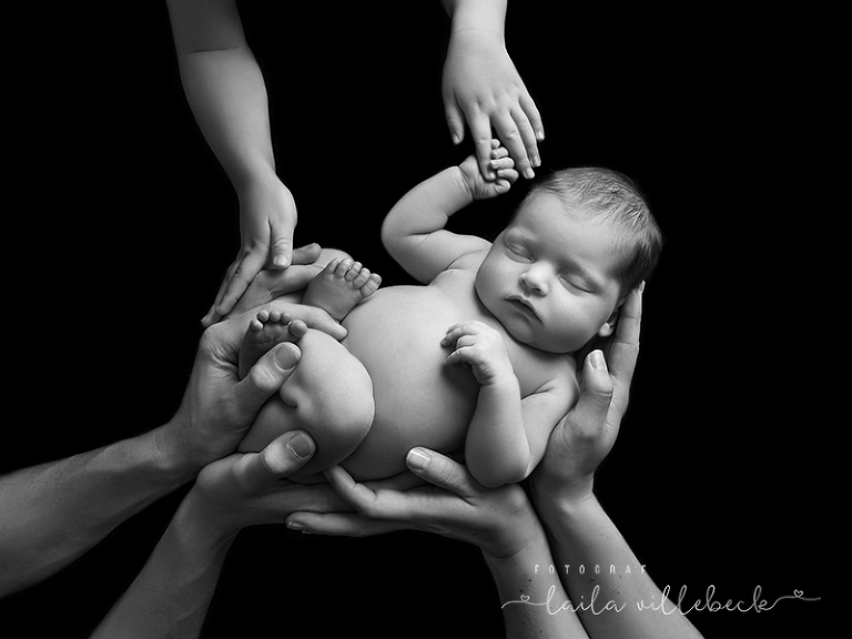Nyfödd bebis på svart bakgrund omgiven av övriga familjemedlemmarnas händer. Svartvitt.