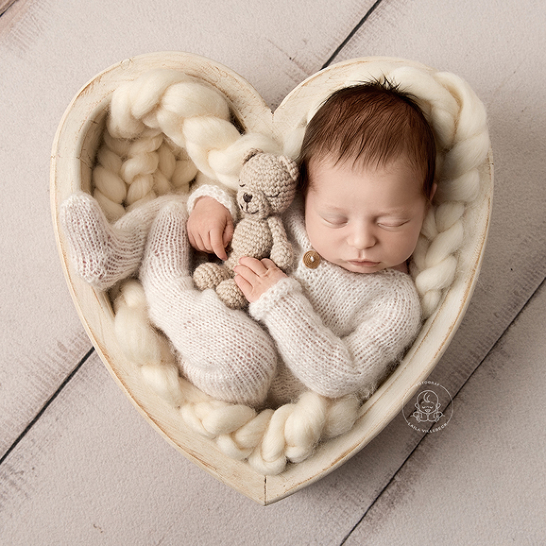 Nyfödda Ossian ligger och sover i en hjärtformad skål. Han kramar om en liten miniatyrnalle och hela bilden går ton i ton i off white.
