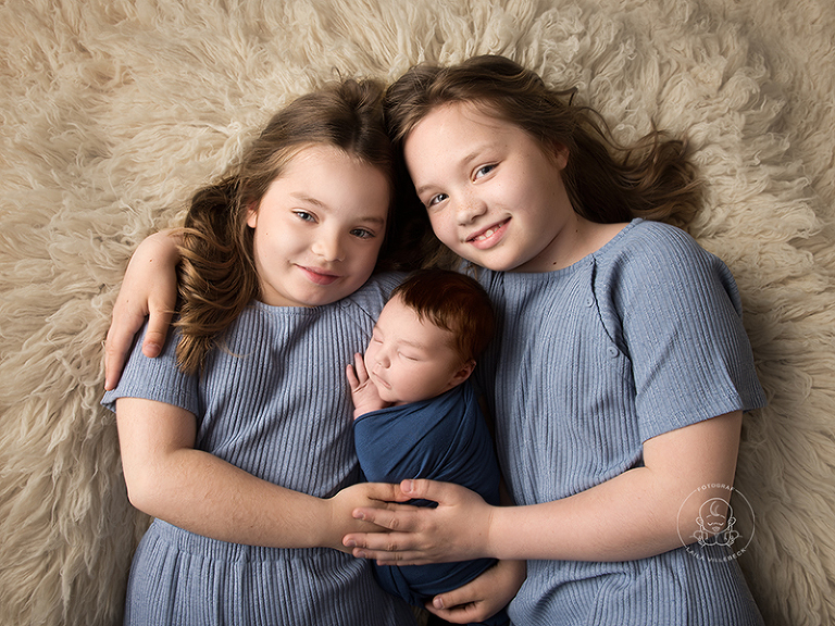 Triss i systrar. Två stolta storasystrar ligger på en fäll och håller sin nyfödda lillasyster emellan sig.