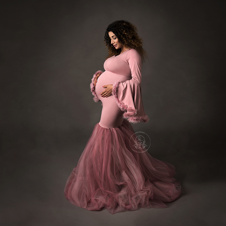 Ett helkroppsporträtt av en gravid kvinna. Hon bär en rosa klänning i sjöjungfrumodell. Klänningen har utsvängda ärmar med pälskant och nederdelen av klänningen är av rosa tyll. Hon står i profil och tittar ner på gravidmagen.