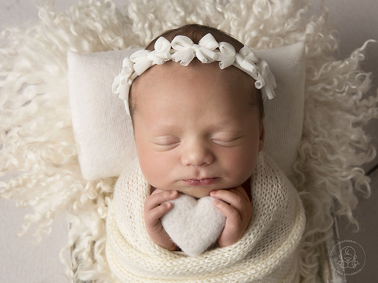 En nyfödd tjej håller ett vitt hjärta med sina händer under hakan. På huvudet har hon ett vitt hårband med rosetter på rad.