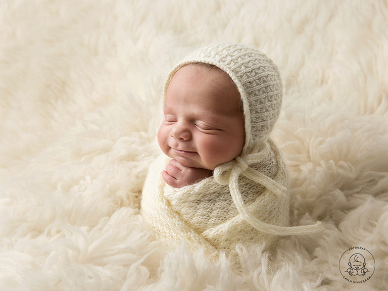 En leende nyfödd bebis är inlindad i vit stickad linda med matchande mössa.