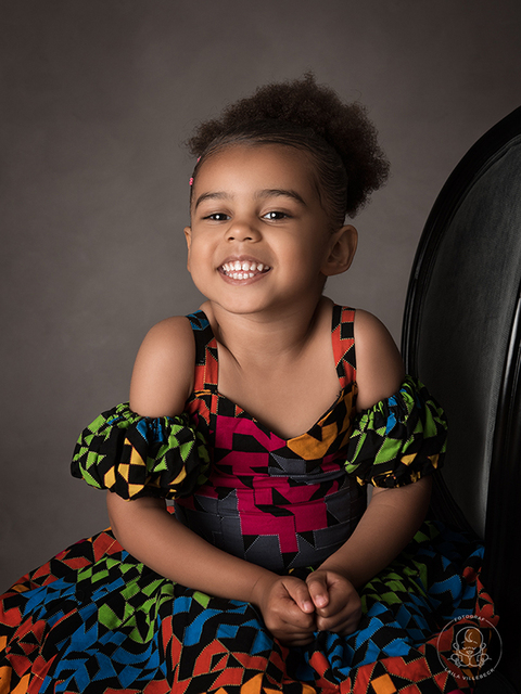 En glad och busig tjej i afrikansk klänning sitter på en stol i studion under en barnfotografering