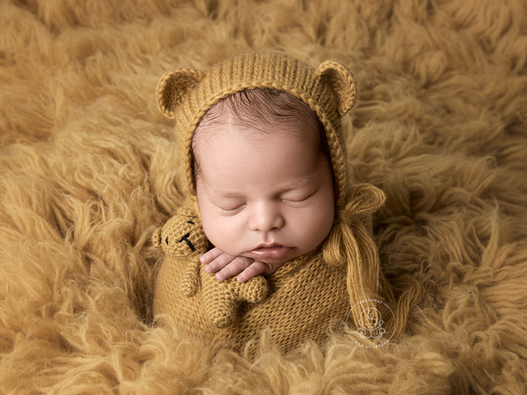 Nyföddfotografering med en inlindad bebis på en senapsgul flokati. Hon har en liten nallemössa på sig och i handen håller hon en nalle i miniatyr.