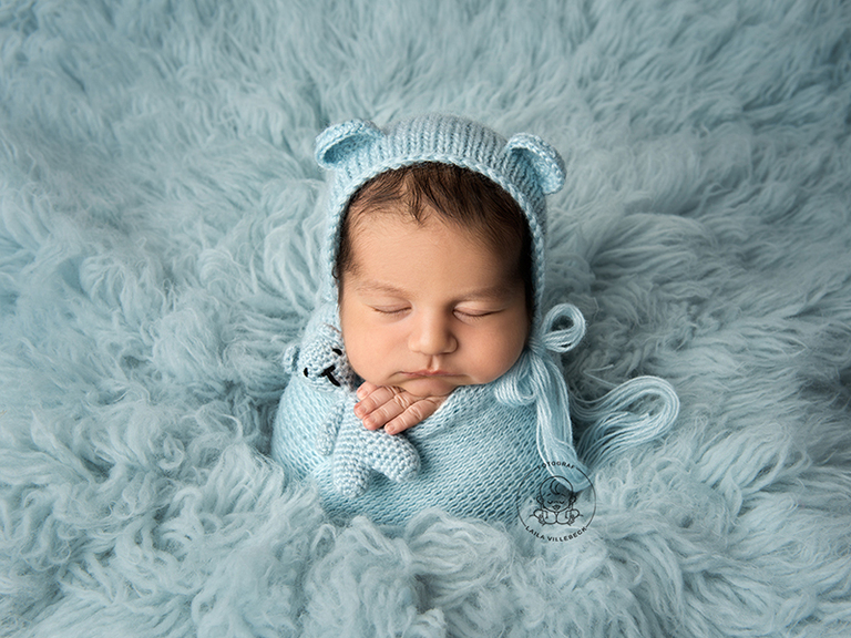 En pojke under sin nyföddfotografering i Linköping. På en ljusblå fäll med matchande stickad nallemössa och teddy i sin hand.