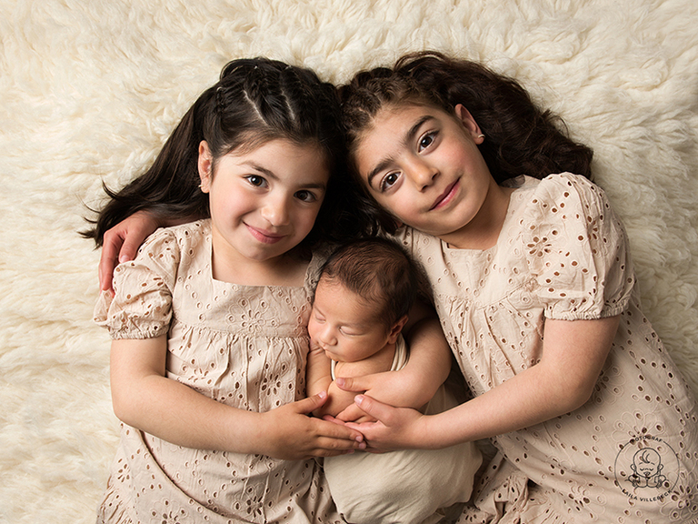 Syskonbild på nyfödd kille med sina två storasystrar. De ligger på en cremefärgad fäll och håller om varandra.