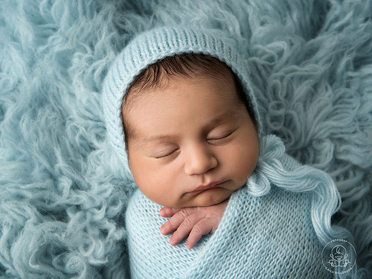 Vacker avskalad bild av nyfödd på en ljusblå fäll, iklädd matchande mössa och inlindad i en stickad wrap.