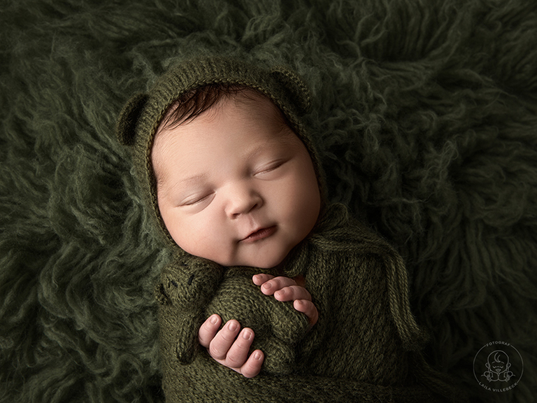 Nyföddfotografering med bebis från Mjölby. Temafärg FOREST. Lilla Eir håller i en nalle och ler ett litet leende.