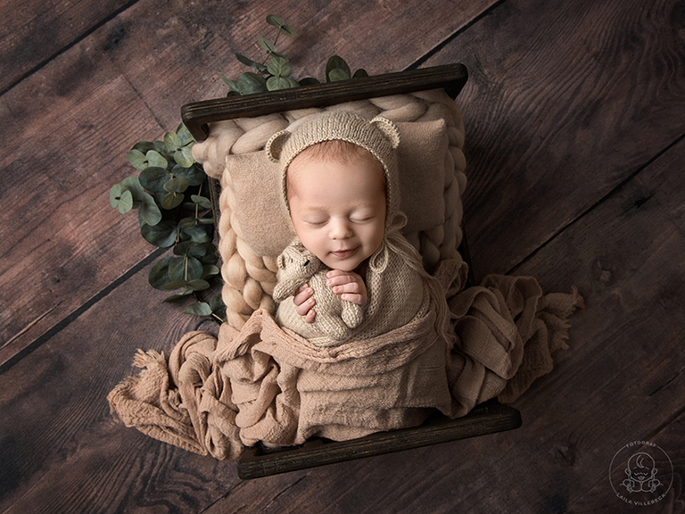 Nyföddfotograf Örebro - nyföddfotografering med Lucas där han ligger i en miniatyrsäng med nallemössa och nalle i händerna
