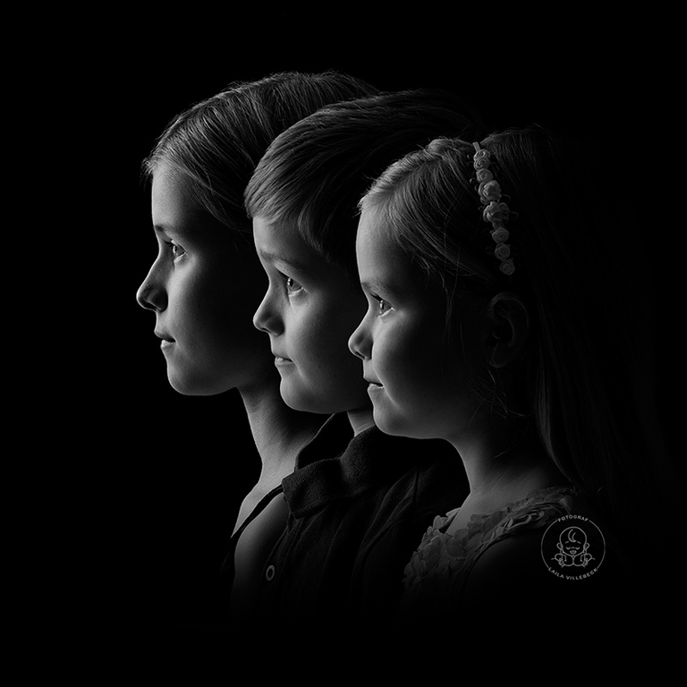 Syskonbild med tre syskon i svartvitt där barnen står i profil i vackert ljus mot svart bakgrund.