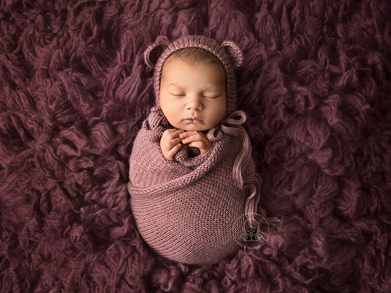 Nyfödd flicka från Nyköping ligger i ett hav av rosa ull, inlindad i matchande stickad wrap och med nallemössa och en liten nalle i händerna.