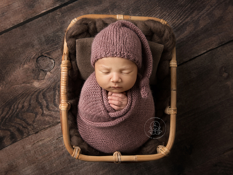 Noelia från Nyköping på nyföddfotografering i Linköping. Här är hon wrappad i en gammelrosa stickad wrap och har en matchande sleeping hat från Dreaming Carita på sig. Hon ligger tryggt i en korg på en mörkt, gammeldags brädgolv.