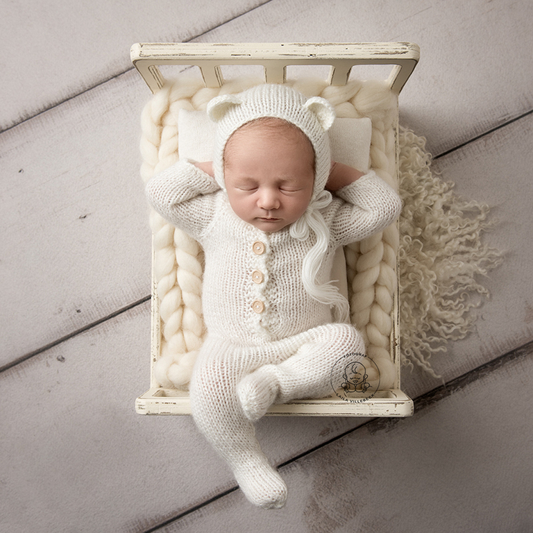 Sötaste nyfödda Eddie ligger i en miniatyrsäng under sin nyföddfotografering hos Fotograf Laila Villebeck. Han har armarna upp och under huvudet och ett av hans ben dinglar avslappnat över kanten på sängen.