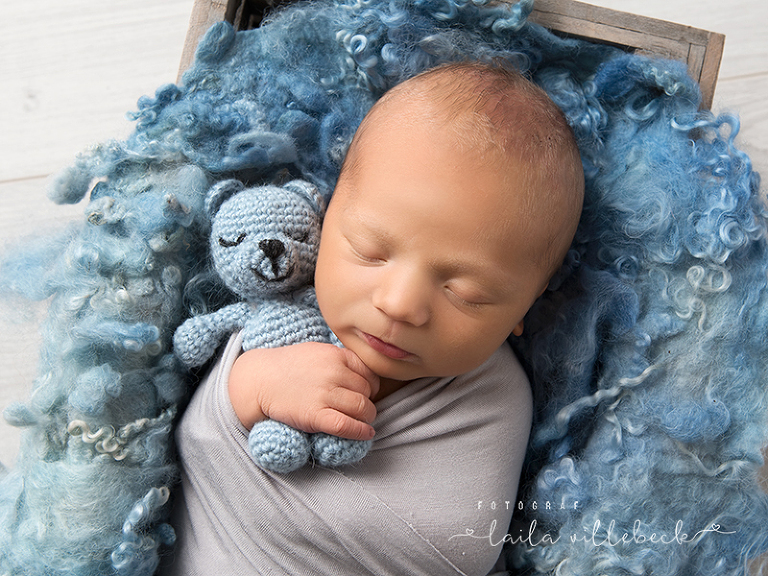 Nyfödd pojke invirad i en grå sjal och sovandes på blå filt med blå nalle i famnen.