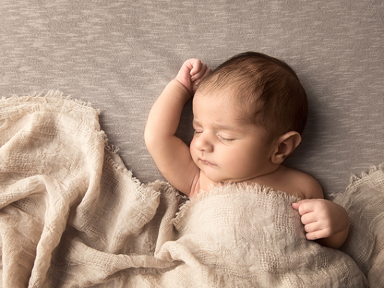 En nyfödd liten pojke ligger och sover under en filt med ena armen sträckt över huvudet