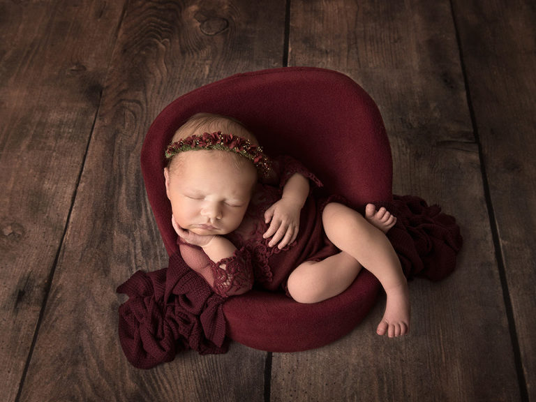 Nyfödd flicka i vinrött sitter och sover i fåtölj under nyföddfotografering