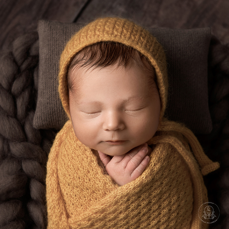 Rofyllt porträtt av en sovande nyfödd pojke iklädd en stickad senapsgul mössa och invirad i en matchande filt.