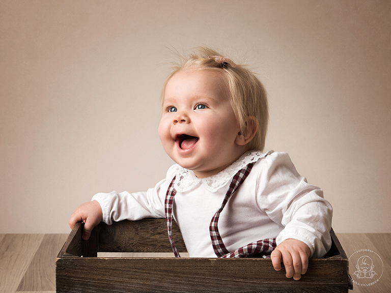 En glad ettåring ler stort när hon sitter i en låda under sin ettårsfotografering.