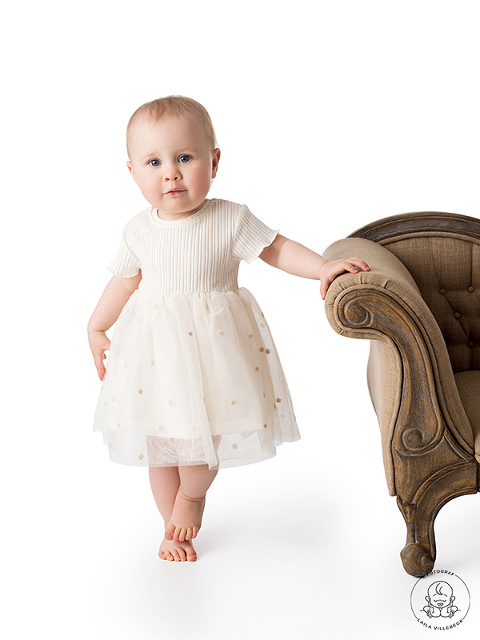 Ettåriga Maja står med ena handen på en soffkant och andra handen på sin fina, vita klänning. Fötterna står lite nätt i kors.