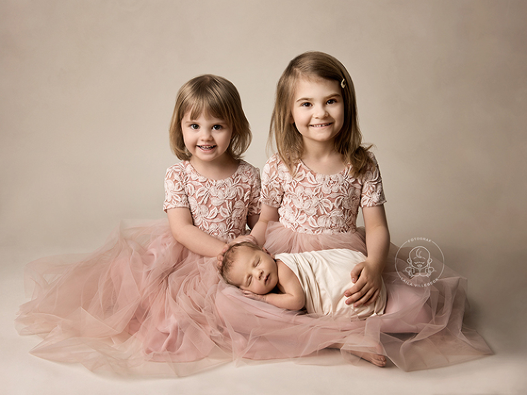 Två söta systrar i likadana rosa tyllklänningar sitter med sin nyfödda lillebror i knät och ler in i kameran