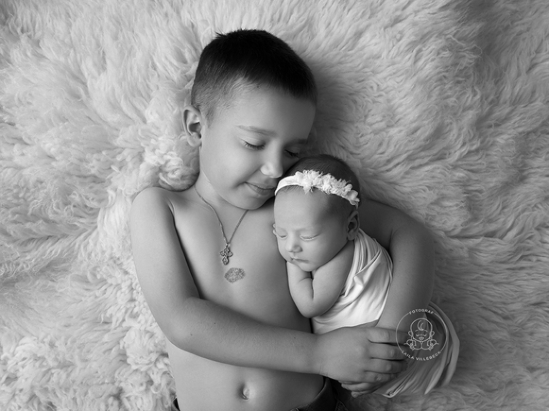 En stolt storebror kramar om sin nyfödda lillasyster. En svartvit bild där barnen ligger på en mjuk, vit fäll. Storebror har ett halsband med kors och lillasyster är inlindad.