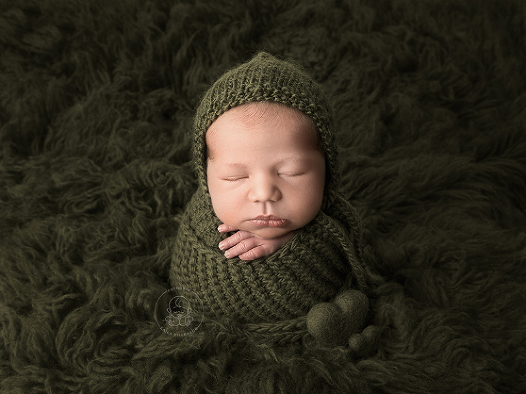 Nyfödd pojke invirad en en härligt murrigt grön stickad filt, placerad på en mörkgrön flokati. Två små hjärtan som går ton i ton med resten av det gröna i bilden pyntar bilden och är placerade i botten av bebisens stickade wrap.