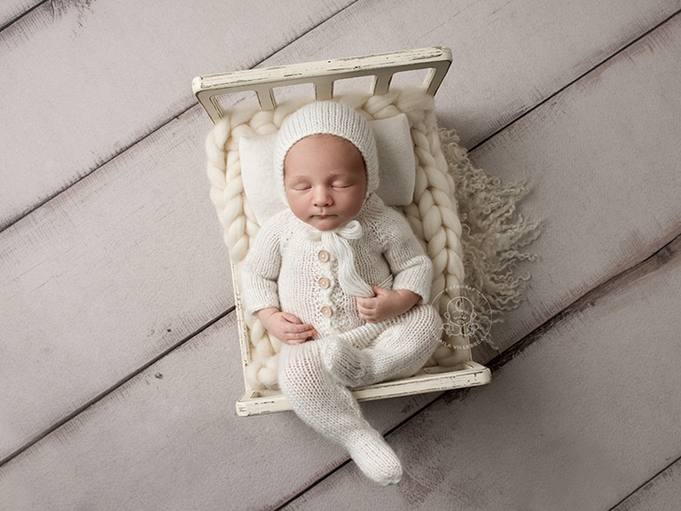 Lille Eddies nyföddfotografering i Linköping. Här ligger han och sover i en vit liten säng iklädd en stickad vit sparkdräkt och matchande mössa.